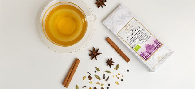 Glas Teetasse mit goldenem Chai-Tee, daneben Verpackung des Ronnefeldt Golden Curcuma Chai und lose Gewürze wie Zimt, Sternanis und Kardamom auf weißem Hintergrund.